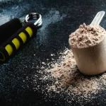 Proteīna pulveris mērvienībā uz galda ar roku trenažieri fonā – piemērots pēc treniņa muskuļu atjaunošanai.