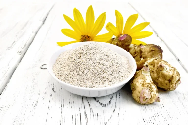 Topinambūru pulveris baltā bļodā ar svaigiem topinambūriem un ziediem uz balta galda