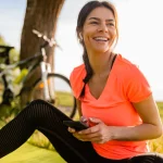 Sieviete sporta tērpā pēc treniņa sēž zālē ar telefonu rokās – veselīgs dzīvesveids dabā