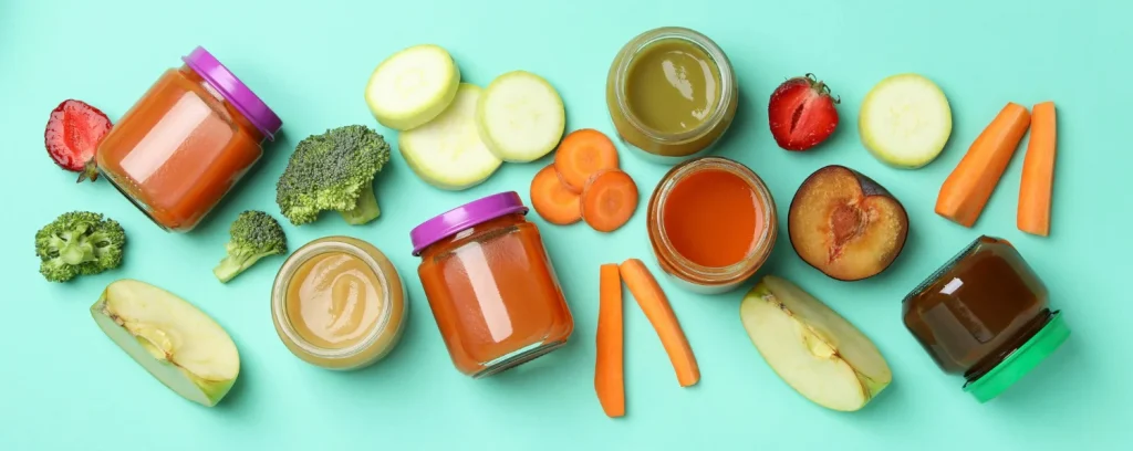 Pārtikas burciņas un svaigi dārzeņi un augļi — brokoļi, burkāni, kabači, āboli, zemenes, plūme uz gaiši zaļa fona.