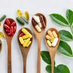 Vitamīnu kapsulas un tabletes koka karotēs ar zaļām lapām uz balta fona