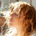 Sieviete ar rudiem, viļņainiem matiem saules gaismā – enerģija un līdzsvars organismā.